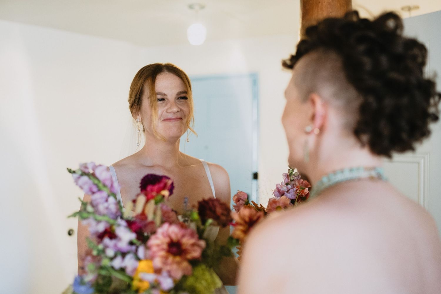 Two brides share a joyful moment, one holding a colorful bridal bouquet of pink and burgundy flowers.