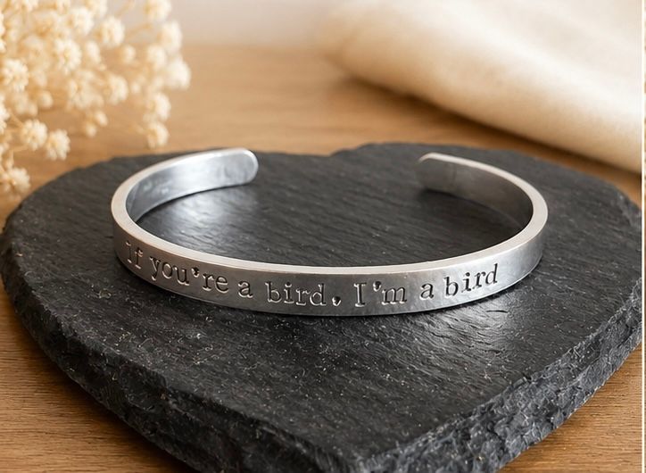 Silver cuff bracelet engraved with 'if you're a bird, I'm a bird' quote, displayed on slate stone.