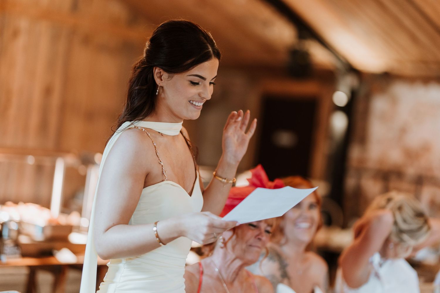 Smiling bride in white dress reads speech at rustic barn wedding reception, waving to guests.