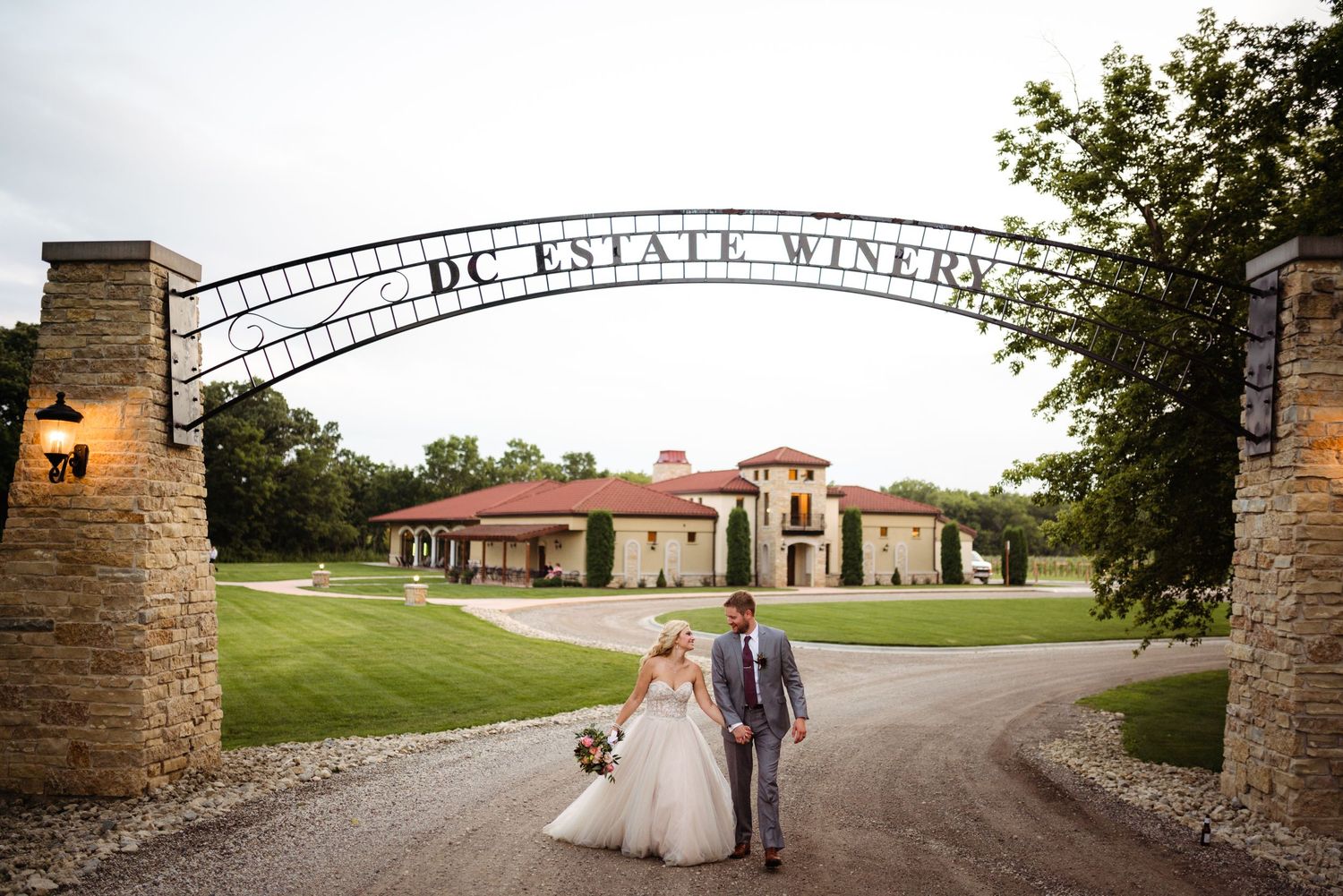 Bride and groom walking under DC Estate Winery iron arch gate with elegant vineyard venue in background.