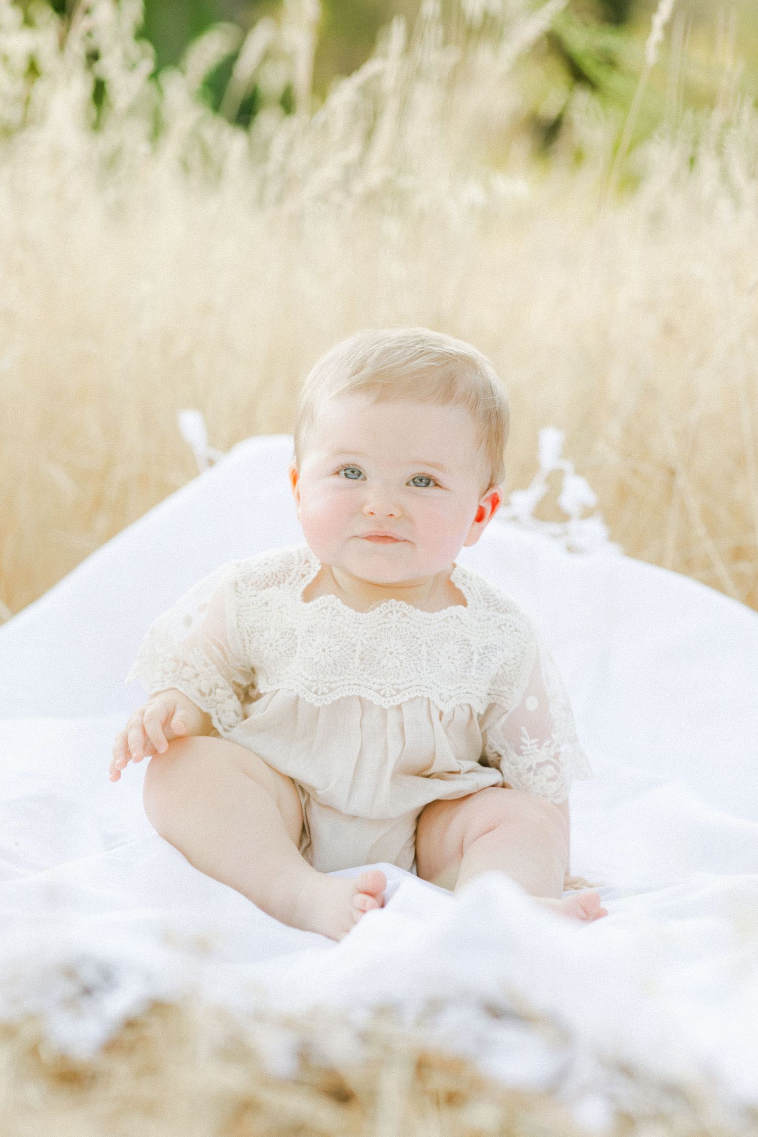 Adorable baby in cream lace romper sitting on white blanket in golden field, looking at camera with blue eyes.