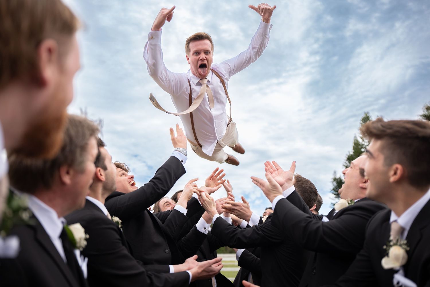 Groom is joyfully tossed into the air by groomsmen in formal attire under cloudy sky.