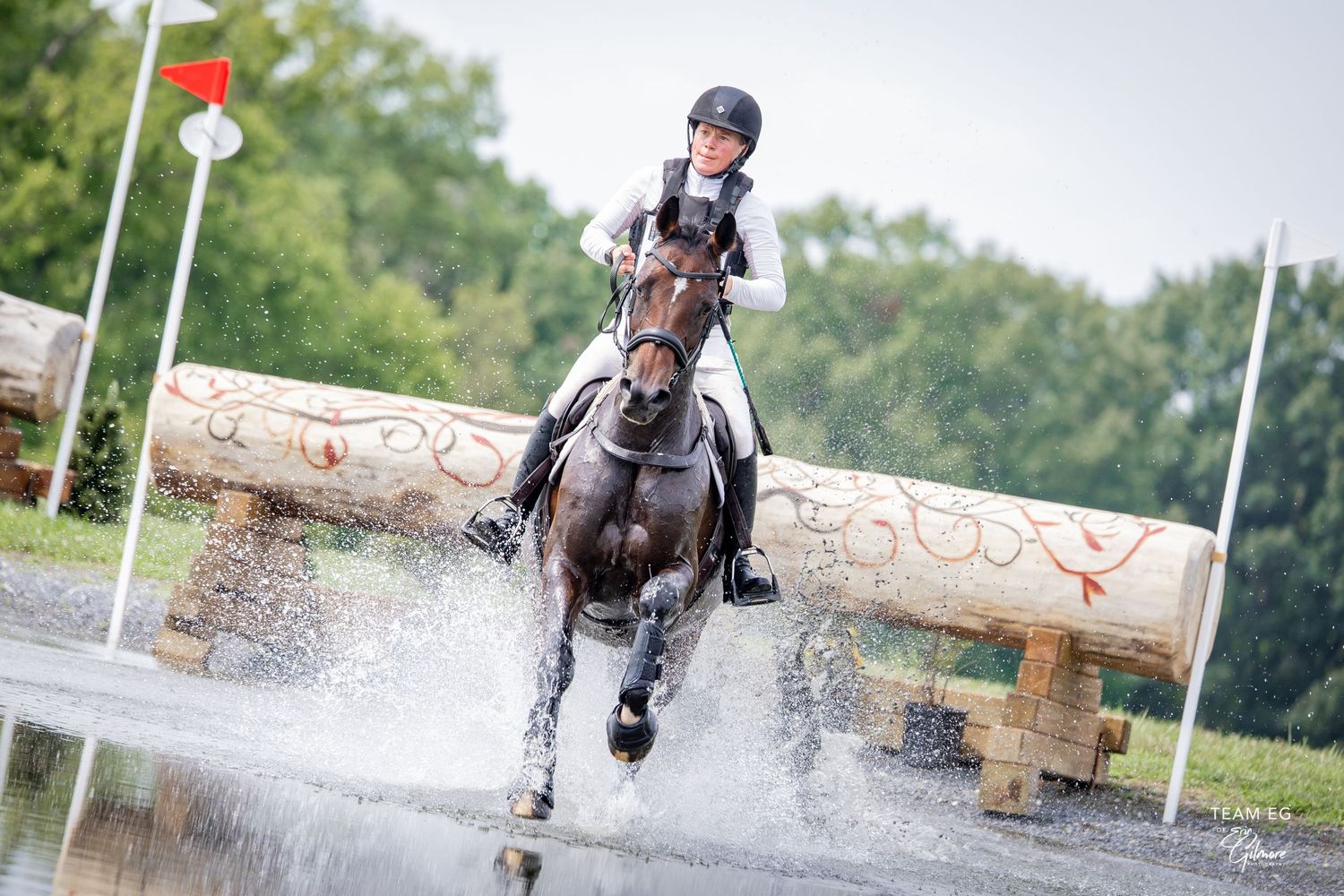 Equestrian rider on dark horse splashing through water obstacle during cross-country competition outdoors.