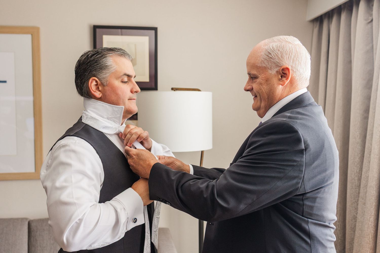 An older man helps adjust the tie of a groom in a vest and white shirt before a wedding ceremony.