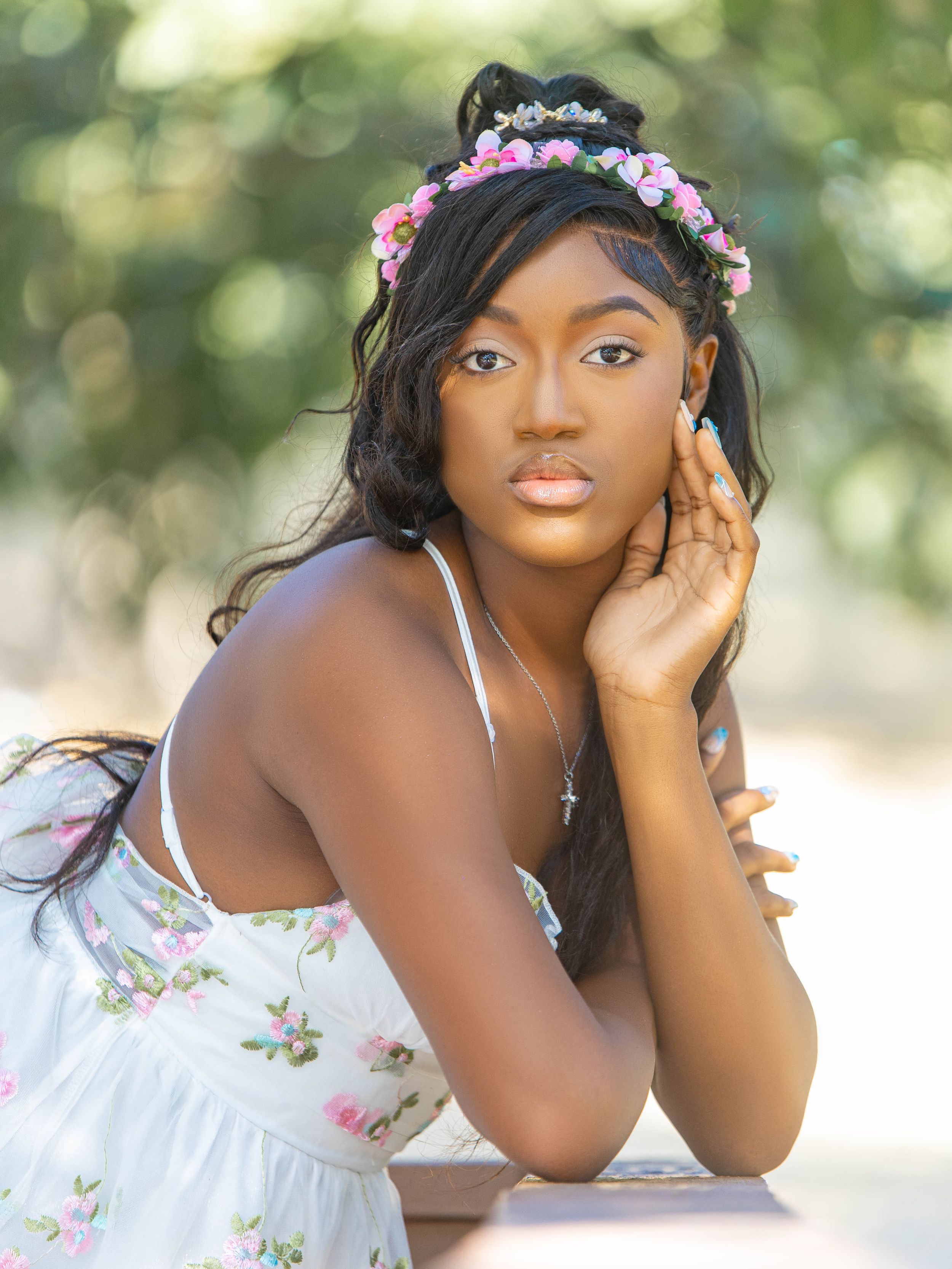 Young Black woman in floral dress and pink flower crown poses gracefully outdoors in soft natural light.