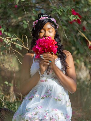 Young woman in floral white dress smelling pink bougainvillea bouquet outdoors, wearing flower crown.