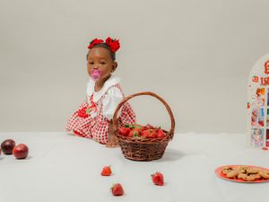 Toddler girl in red gingham dress with bow, sitting behind a wicker basket of strawberries on a white table.