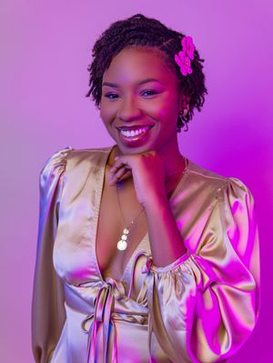 Smiling young woman in gold satin dress with pink flower hair accessory poses against purple background.