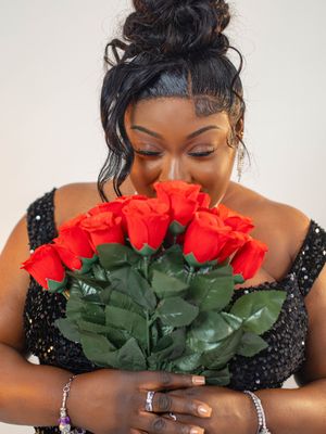 A woman in a black sequin dress smells a bouquet of red roses, eyes closed, wearing bracelets.