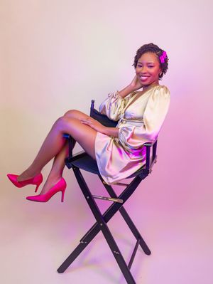 A smiling woman in a satin dress and pink heels sits gracefully on a director's chair against a pink backdrop.