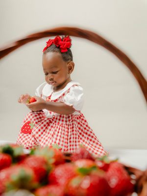 A toddler girl in a red gingham strawberry dress holds a strawberry near a basket of fresh strawberries.