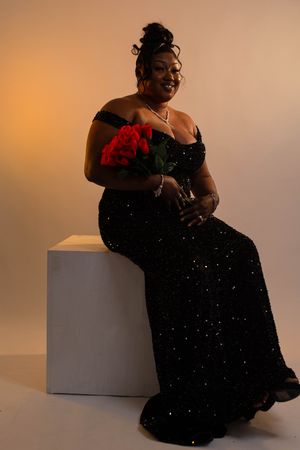 Woman in black sequin off-shoulder gown holding red roses, seated on white cube in elegant studio portrait.