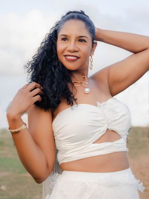 Woman in white crop top and skirt with gold jewelry smiling outdoors, curly black hair, pearl necklace.