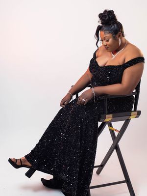 A woman in a black sequin off-shoulder gown sits gracefully on a director's chair against a white background.