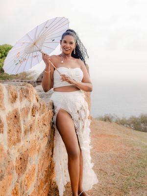 Woman in white crop top and skirt holding a parasol, leaning against a stone wall with ocean views.