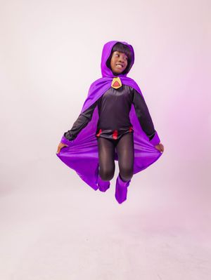 Person in purple superhero cape and hood costume jumping mid-air against light background.