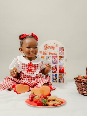 Toddler girl in red gingham dress celebrates 1st birthday with strawberries and cookies beside a monthly photo display.