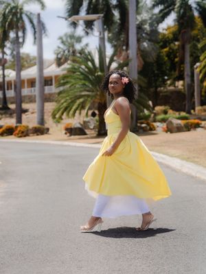 Woman in yellow midi dress and white pants walks confidently on sunny road lined with palm trees.