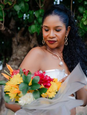 Bride in white off-shoulder gown holding vibrant tropical bouquet with yellow and red flowers, wearing pearl necklace and earrings.
