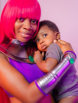 Woman in pink wig and purple costume holding a baby in gray shirt, posing for a cosplay photo shoot.