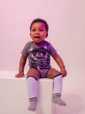 Adorable baby boy sitting on white surface wearing gray graphic onesie and white knee-high socks.