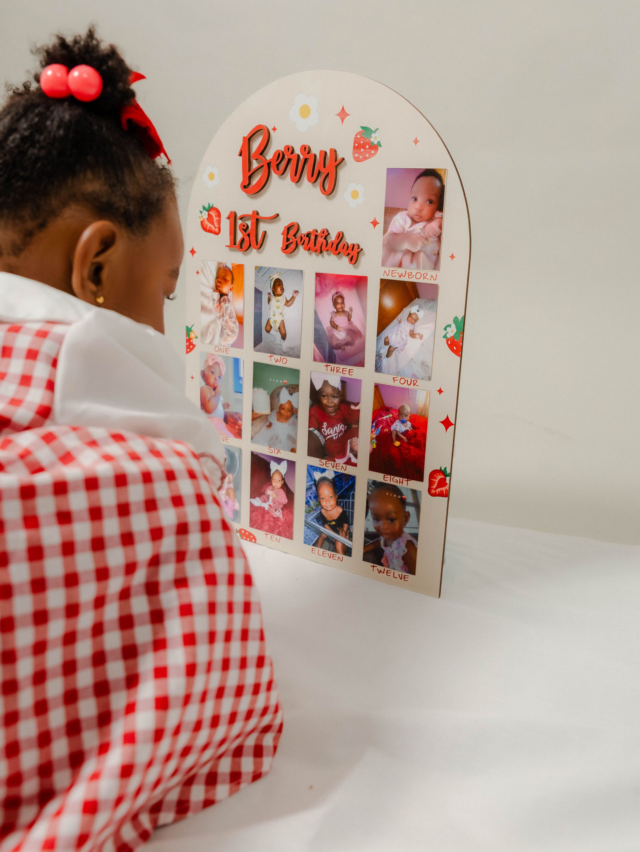 A toddler girl in red gingham dress views her first birthday photo collage board named Berry's 1st Birthday.