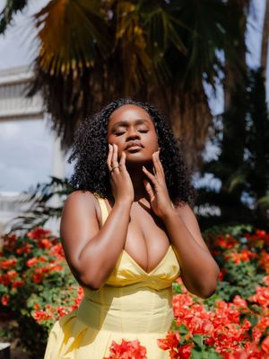 A woman in a yellow dress poses gracefully among red flowers with a palm tree backdrop.