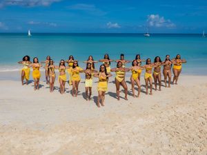 Group of women in matching yellow bikinis forming a heart shape on a tropical white sand beach.