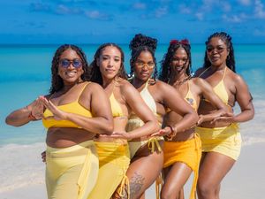 Five women in matching yellow swimsuits pose together on a tropical white sand beach with turquoise water.