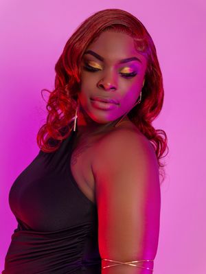A Black woman in a black dress with bold yellow eye makeup and red wavy hair poses against a pink-purple background.