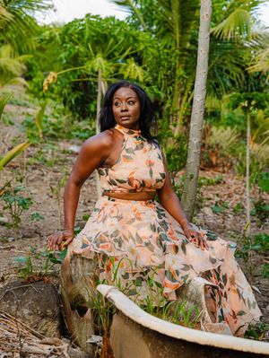 Woman in floral halter crop top and skirt sits on vintage bathtub surrounded by tropical palm trees.