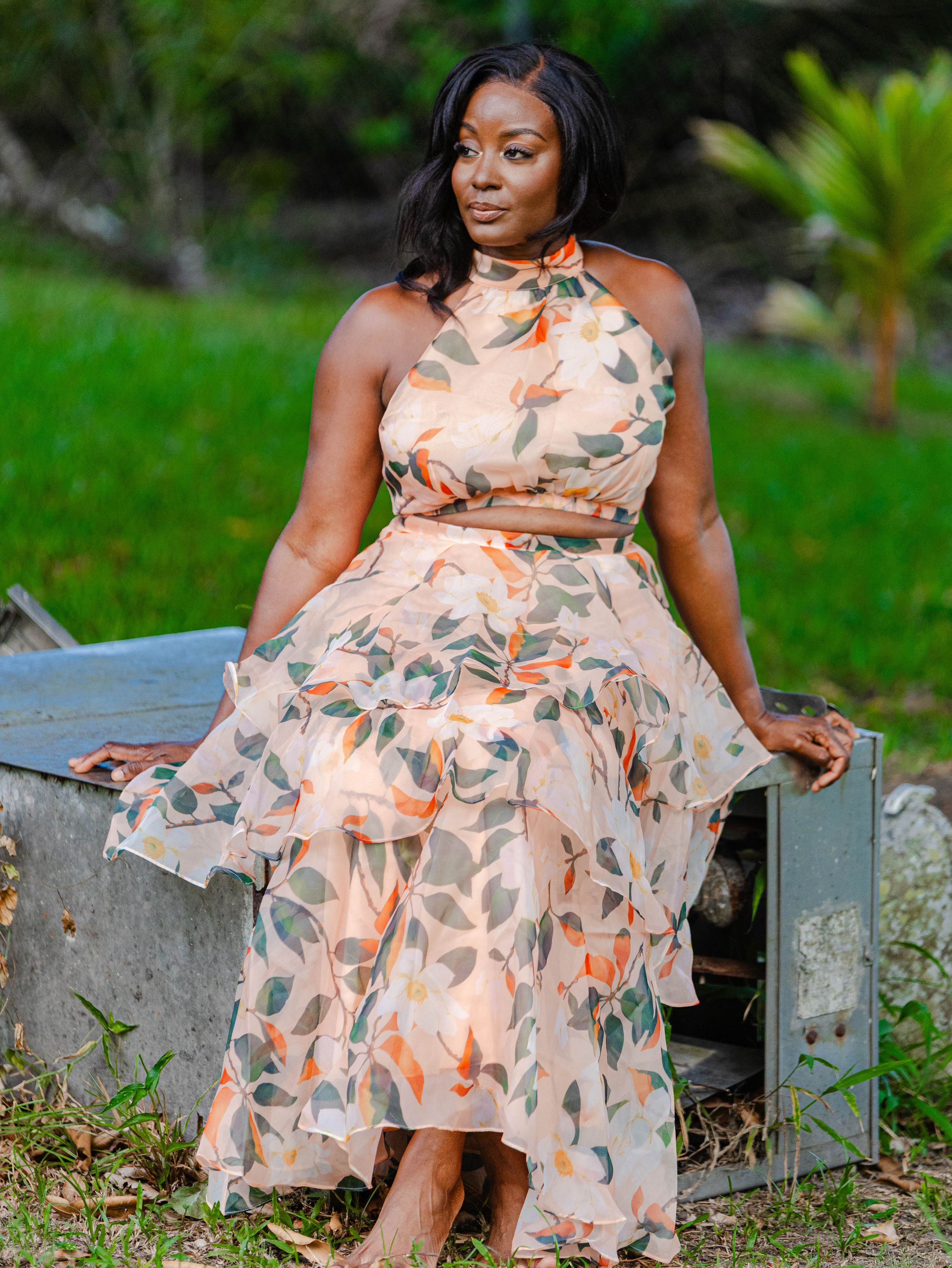 Woman in floral halter midi dress sitting outdoors in lush green garden setting.