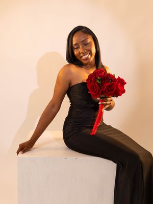 Woman in black strapless gown sitting on white cube holding red roses bouquet, smiling elegantly in studio setting.