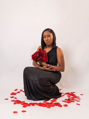 Woman in black dress sitting on floor holding red roses bouquet surrounded by scattered rose petals on white background.
