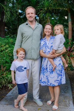 Family of four posing outdoors among tropical greenery, parents with two young red-haired children in summer attire.