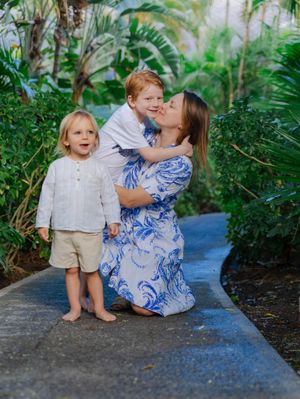 A mother in a blue floral dress hugs her redhead child outdoors while a young blonde toddler stands nearby.