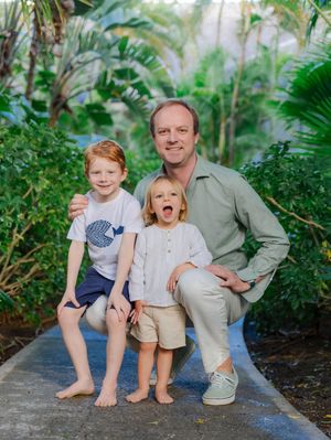 A man crouches with two young children on a garden path surrounded by lush tropical greenery.