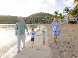 Family of four walking hand-in-hand on a tropical beach at sunset with palm trees and green hills in the background.