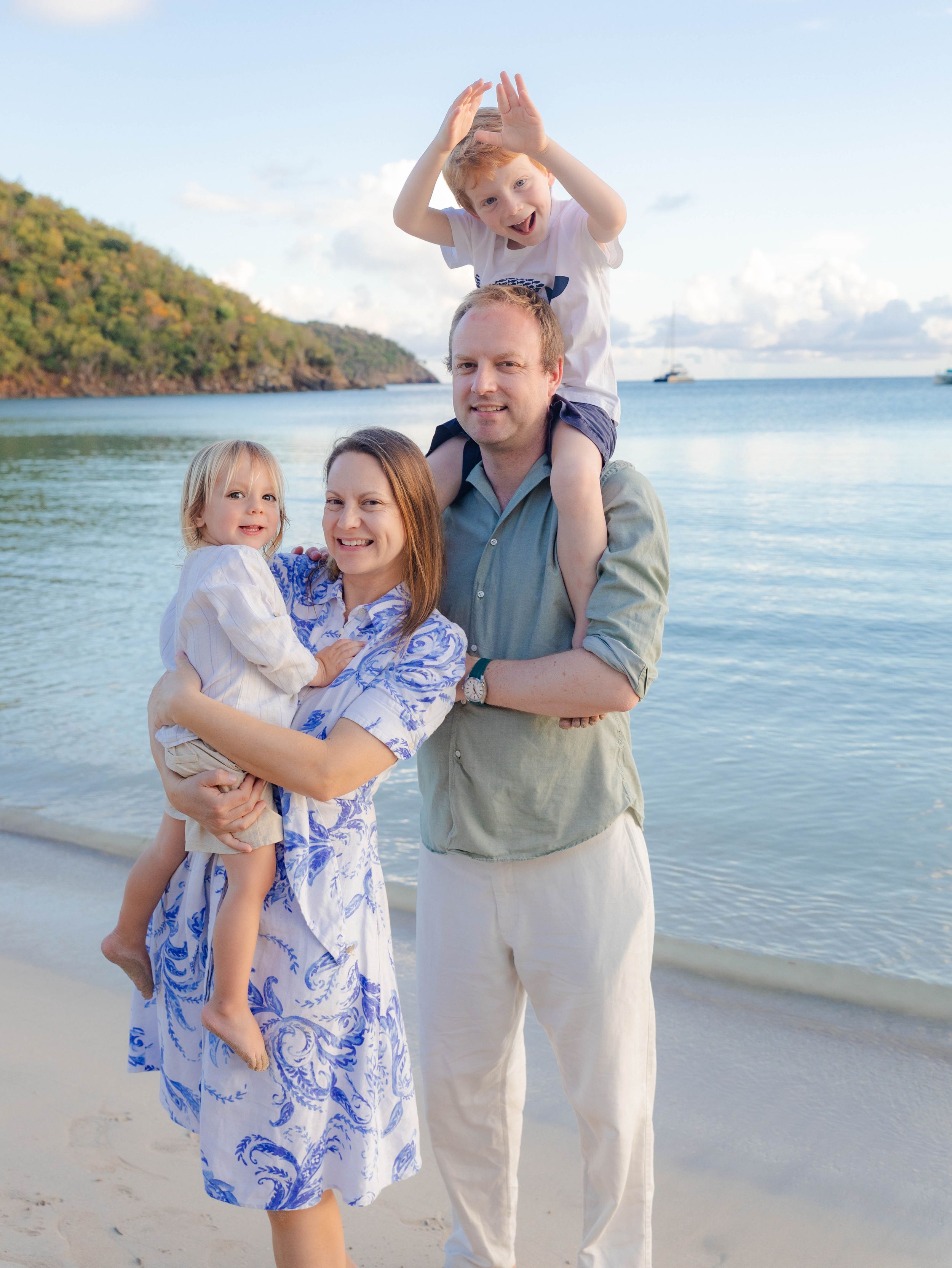 Happy family of four posing on a tropical beach, parents holding young children with turquoise water in background.