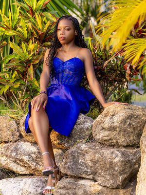Young woman in royal blue lace dress posed on rocks surrounded by tropical foliage.