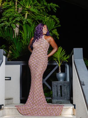 Woman in pink floral mermaid gown with purple hair poses on steps surrounded by tropical greenery.