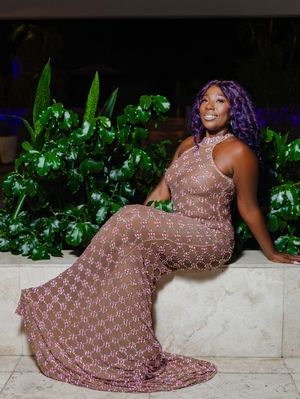 Woman in purple hair wearing elegant pink lace gown poses gracefully beside lush tropical greenery at night.