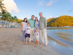 Family of six poses on a tropical beach with palm trees and lush green hills in the background.