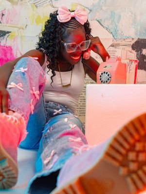 Young woman in pink bow, white top, bow-print jeans, and colorful Timberlands sits against a vibrant collage wall.
