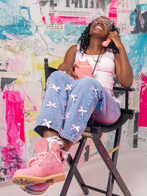 Young woman in pink bow, white tank, bow-print jeans, and pink Timberlands sits in director's chair by collage wall.