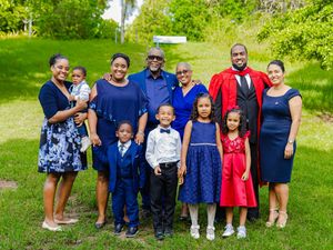 Family group photo outdoors in formal attire, including a graduate in red gown, children, and adults on green grass.