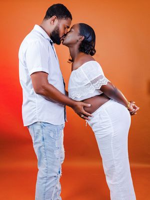 A couple in matching white outfits share a kiss while the woman cradles her baby bump against an orange background.