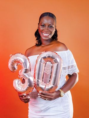 A smiling woman in a white off-shoulder dress holds rose gold number 30 balloons against an orange background.
