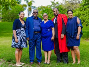 Family of five posing outdoors in blue outfits with one member in graduation regalia on a green lawn.
