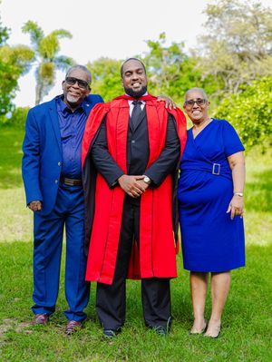 Graduate in red academic robes poses with proud parents in blue outfits outdoors on green lawn.